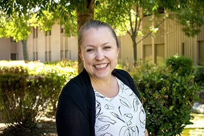 headshot of a blonde Caucasian female in a black and white patterned shirt and black cardigan against a nature background